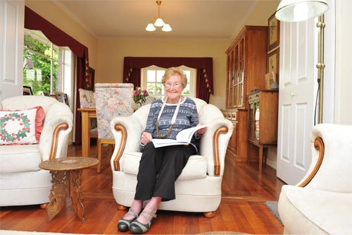 Joan sits in an armchair with a book on her lap at home