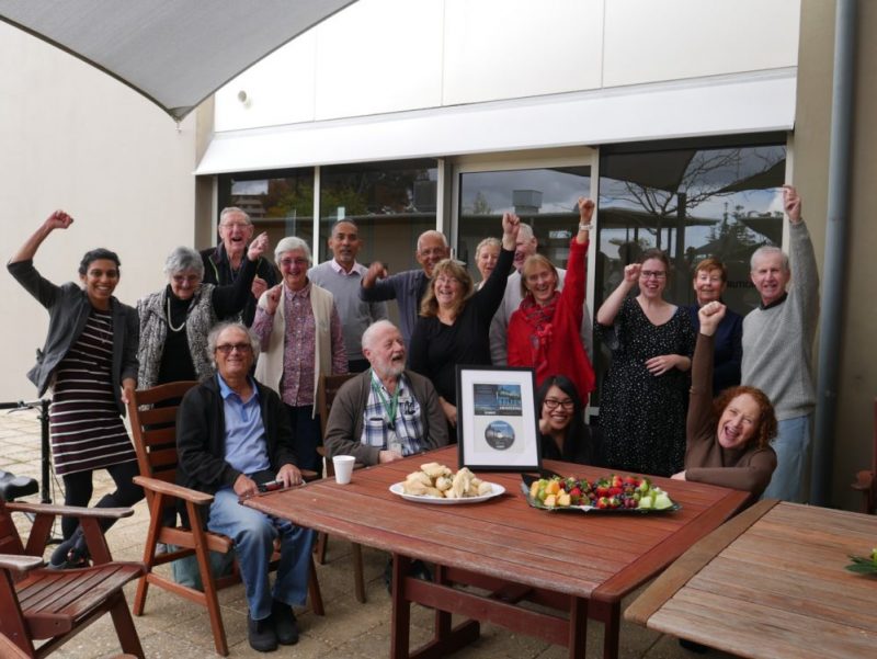 Seventeen people gathered around a table, punching the air with happiness. Table has food and certificate on display.