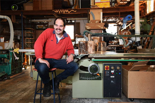 Man sitting in his woodwork studio surrounded by his artwork, smiling at the camera