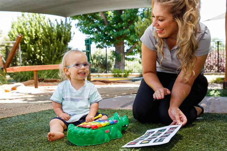 A young boy plays on the grass with a xylophone, and VisAbility team member Caris holds an aided language display