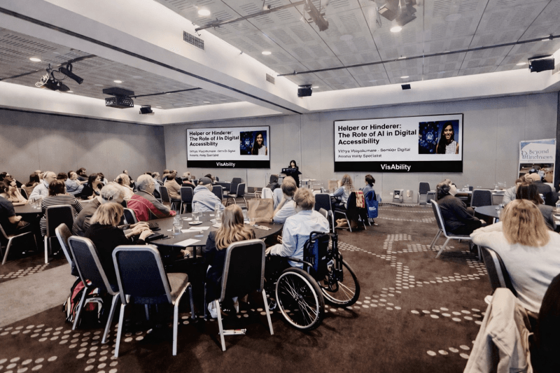 A room full of people sitting at tables look up to watch Vithya Vijayakumare stand at a podium and present at the Round Table on Information Access for People with Print Disabilities Conference